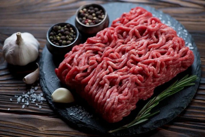 Raw lean ground beef displayed on a stone surface with salt, pepper, and a slice of garlic surrounding it.
